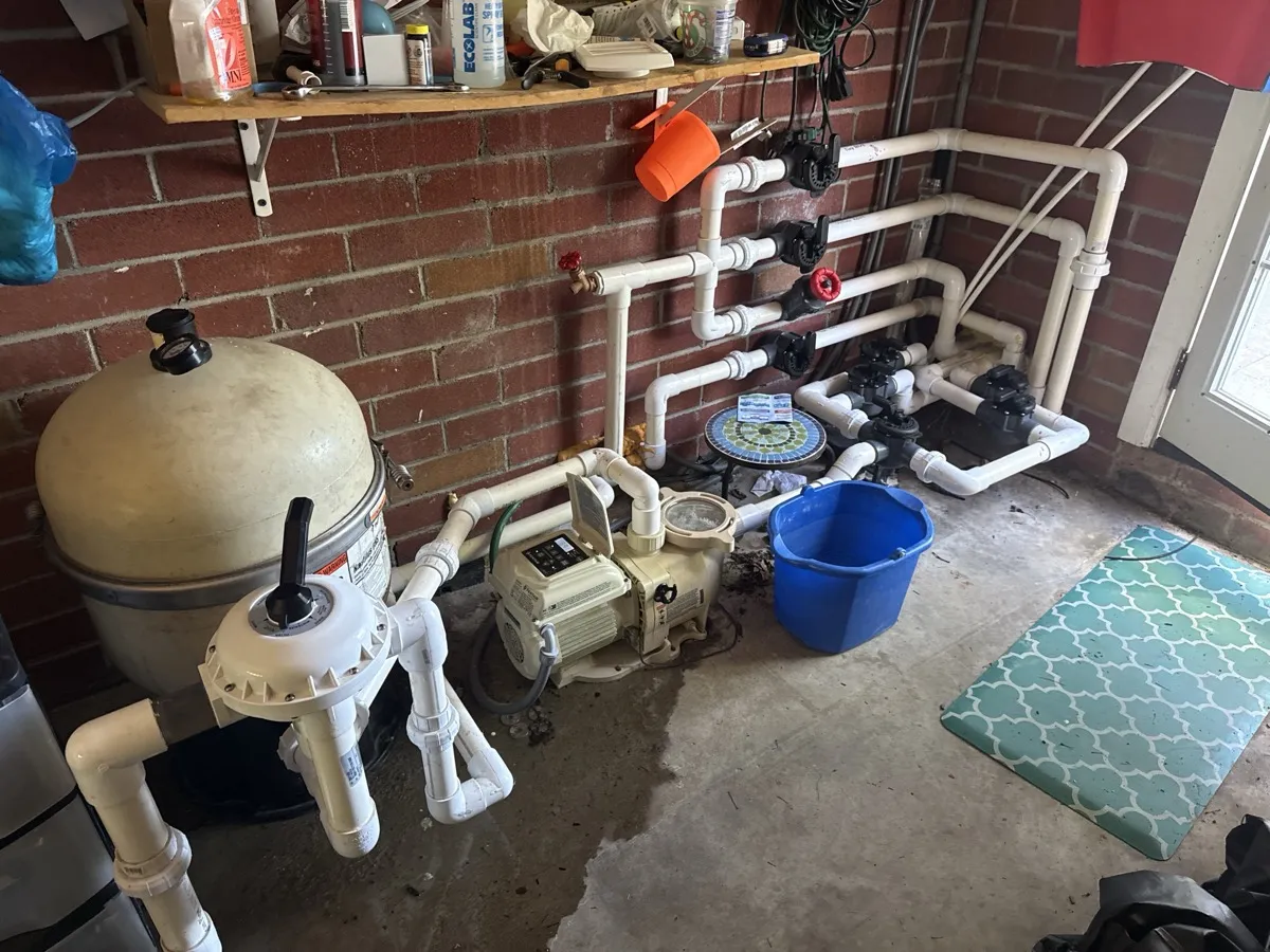 A real backyard pool equipment pad: filter tank, pump, and a wall of PVC valves against a brick wall